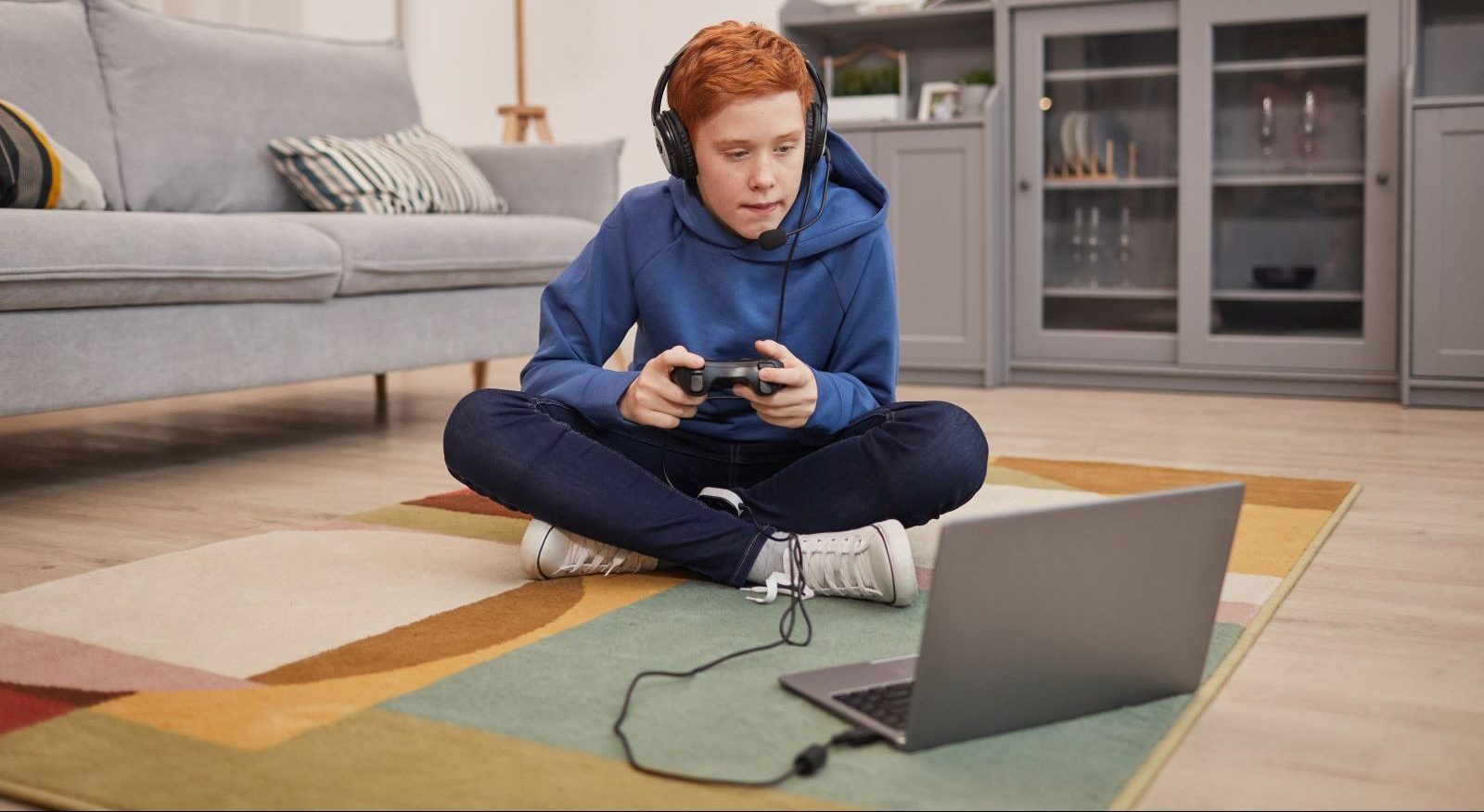Teen sits on living room floor playing video games on a laptop.
